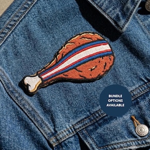 May include: An embroidered patch shaped like a fried chicken leg with a golden-brown crust and a white bone end. A horizontal stripe featuring red, white, and blue colors runs across the center of the chicken leg. The patch is attached to denim.