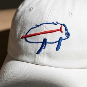May include: A white baseball cap featuring an embroidered graphic on the front panel. The design consists of a blue outlined shape resembling a stylized bison, accented with a horizontal red line running through the center of the figure.