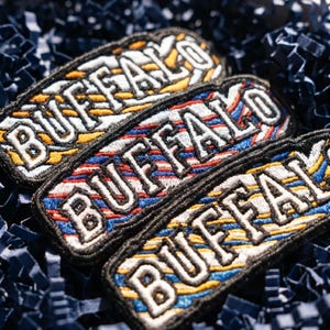 May include: Three rectangular embroidered patches featuring the word BUFFALO in bold white capital letters with a black border. Each patch displays a distinct background pattern of diagonal stripes in varying color combinations of blue, red, and yellow.