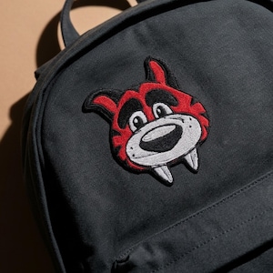 May include: A charcoal gray backpack featuring an embroidered patch of a cartoon tiger head. The tiger is rendered in red, black, and white thread, showcasing prominent ears, thick eyebrows, a wide snout, and two visible lower fangs on its face.