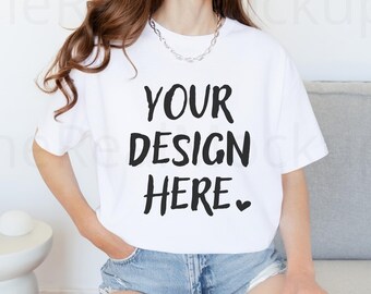 Puede incluir: Una camiseta blanca con el texto "YOUR DESIGN HERE" en negro, combinada con pantalones cortos de mezclilla. La modelo lleva un collar de cadena plateada. La camiseta es una maqueta, lista para diseños personalizados.
