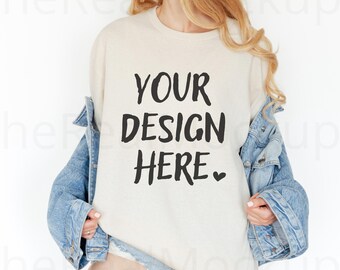 May include: Cream-colored sweatshirt with the text "YOUR DESIGN HERE" in black. The sweatshirt is worn with a blue denim jacket. The image is a mockup, ideal for showcasing custom designs on apparel.