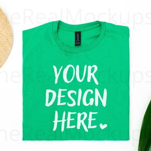 以下が含まれることがあります： 白文字で「YOUR DESIGN HERE」と書かれた、折りたたまれた緑色のTシャツ。麦わら帽子と緑の葉が画像の一部に写っています。Tシャツはカスタムデザインのモックアップです。