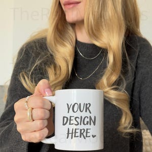 Puede incluir: Taza de cerámica blanca con el texto "YOUR DESIGN HERE" en negro. La taza se sostiene frente a una persona que lleva un suéter gris. La taza es de tamaño estándar para café o té.