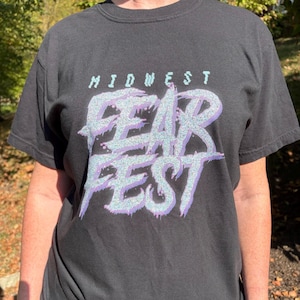 Peut inclure: T-shirt noir avec l'inscription "MIDWEST FEAR FEST" dans un design scintillant violet et turquoise. Le t-shirt à manches courtes semble être fait d'une matière douce.