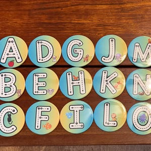 May include: A collection of round, colorful alphabet magnets. Each magnet features a letter of the alphabet with a dotted line for tracing, and a small illustration. The background gradient transitions from yellow to blue.