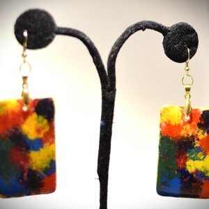 May include: A pair of rectangular earrings with a colorful abstract design. The earrings feature a gold-colored hook and a pendant with a mix of red, yellow, blue, and green hues. The earrings are displayed on a black earring stand.