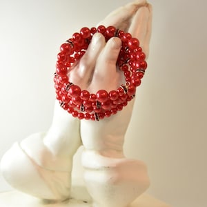 May include: A red beaded bracelet with multiple strands, featuring varying sizes of round beads. The bracelet is accented with small, silver-toned spacers. The jewelry is displayed on a white, sculpted hand.