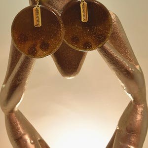 May include: A pair of gold-toned earrings with a circular design. The earrings are a dark brown color with glitter and the word "BEAUTIFUL" on a small gold tag. The earrings are displayed on a gold-colored sculpture.