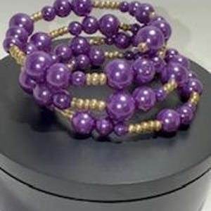 May include: Three beaded bracelets. Each bracelet features large, round, purple beads interspersed with smaller gold-colored beads. The bracelets are stacked on top of each other, showcasing their design and color contrast.