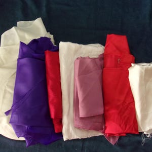 May include: A collection of folded fabric in various colors, including white, purple, red, and pink. The fabrics appear to be silk or satin, with a shiny texture. A pair of scissors is visible in the background.