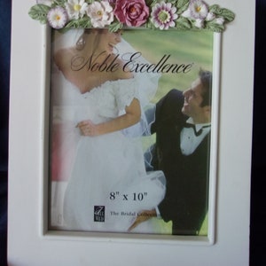 May include: White picture frame with a floral design around the top edge. The frame is for an 8x10 inch photo and has the words "Noble Excellence" and "The Bridal Collection" printed on it.
