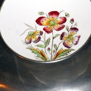 Pu&ograve; includere: Un piatto d'argento con un inserto in porcellana bianca con un motivo floreale con fiori rossi, gialli e verdi. Il piatto ha il numero "181" inciso sul fondo.