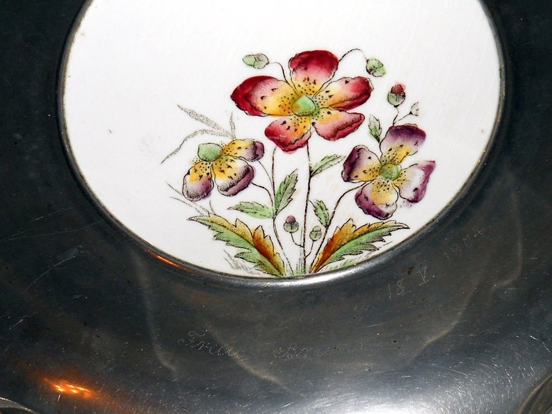 Pu&ograve; includere: Un piatto d'argento con un inserto in porcellana bianca con un motivo floreale con fiori rossi, gialli e verdi. Il piatto ha il numero "181" inciso sul fondo.