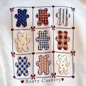 May include: White sweatshirt with a blue and red patchwork design of bears. The bears are arranged in a grid pattern and have different patterns on their bodies. The text "Beary Country" is printed below the bears.