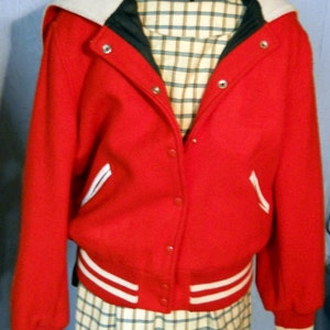 May include: A red letterman jacket with white accents, a cream-colored collar, and a plaid lining. The jacket has white stripes on the cuffs and waist. The jacket is buttoned up and displayed on a mannequin.