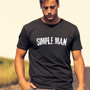May include: A black short-sleeved t-shirt featuring the text "SIMPLE MAN" printed in white across the chest in a distressed, capitalized font. The garment has a standard crew neckline and a straight hemline, providing a basic and functional design.