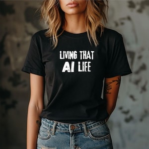 Op de afbeelding: Een zwart T-shirt met korte mouwen en de witte, verweerde tekst "LIVING THAT AI LIFE" op de borst gedrukt. Het shirt wordt gecombineerd met een blauwe spijkerbroek met een zichtbare metalen knoop en stiksels bij de zakken.