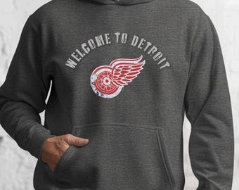 Peut inclure: Sweat à capuche gris foncé avec une poche avant et une capuche. Le sweat-shirt affiche le texte "WELCOME TO DETROIT" au-dessus d'un logo vieilli d'une roue rouge et blanche avec des ailes rouges.
