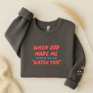 Puede incluir: Sudadera gris oscuro con el texto "WHEN GOD MADE ME HE SMILED AND SAID 'WATCH THIS'" en rojo y azul. La sudadera tiene cuello redondo y mangas largas con puños enrollados.