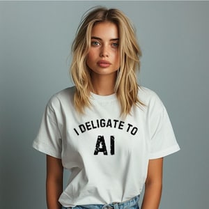 May include: A white short-sleeved t-shirt featuring the black text I DELIGATE TO AI printed in a distressed, bold font across the chest. The shirt has a classic crew neckline and a standard fit, made of a plain white fabric.