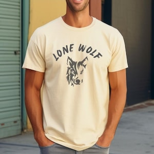 May include: Cream-colored t-shirt with the words "LONE WOLF" arched above a stylized wolf head graphic. The graphic is in dark gray. The shirt has short sleeves and a crew neck.