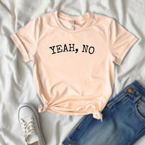 May include: A peach-coloured t-shirt with the text "YEAH, NO" in black. The shirt is knotted at the waist and paired with blue jeans and white trainers. The casual outfit is laid out on a white sheet.