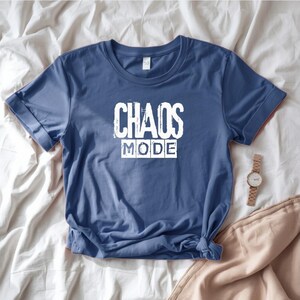 May include: A blue t-shirt with the words "CHAOS MODE" printed in white. The shirt is folded and lies on a white sheet. A gold watch and tan pants are also visible.