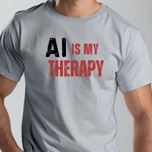 May include: A light grey short-sleeved t-shirt featuring the text AI IS MY THERAPY printed on the chest. The letters AI are in black, while the words IS MY THERAPY are in red. The text has a distressed, weathered texture throughout the design.