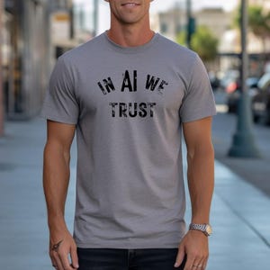 Könnte beinhalten: Ein graumeliertes Kurzarm-T-Shirt mit dem schwarzen Schriftzug "IN AI WE TRUST", der in einer abgenutzten Schablonenschrift auf der Brust gedruckt ist. Das T-Shirt hat einen Rundhalsausschnitt und eine Standardpassform aus strukturiertem Stoff.