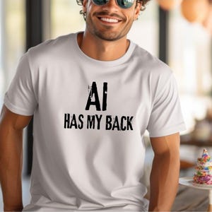 Puede incluir: Una camiseta blanca de manga corta con el texto negro AI HAS MY BACK impreso en el pecho. Las letras tienen un aspecto desgastado y texturizado. La camiseta tiene cuello redondo y un corte estándar, confeccionada en tela blanca lisa.