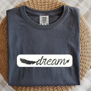 May include: A dark gray t-shirt with a white rectangular graphic. The graphic features a black feather and the word "dream" in a handwritten style font. The shirt is folded neatly on a woven surface.