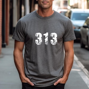 Puede incluir: Camiseta gris oscuro con el número "313" impreso en blanco en el pecho. Los números tienen un aspecto desgastado y texturizado. La camiseta tiene mangas cortas y cuello redondo.