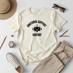 May include: A cream-colored short-sleeve t-shirt featuring a black graphic of a robot head with a headset and the text "EMOTIONAL SUPPORT CHATBOT" printed in a distressed font across the chest area of the garment.