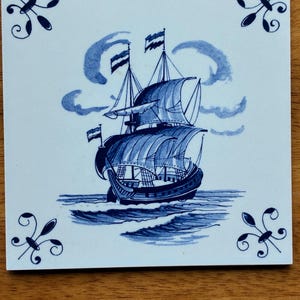 May include: Square ceramic tile with a blue and white nautical design. The image features a sailing ship with billowing sails on a wavy sea, under a cloudy sky. Decorative floral motifs are in each corner.