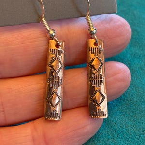 May include: A pair of copper-colored earrings with a rectangular shape and intricate geometric patterns. The earrings have a hook closure and a small silver bead detail. The design features diamond and line patterns, creating a textured look.
