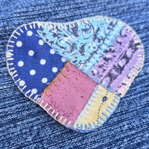 May include: Close-up of a heart-shaped patch sewn onto blue denim fabric. The patch is made of various fabric pieces in blue polka dots, patterned blue, purple, pink, and yellow. The patch is outlined with white stitching.