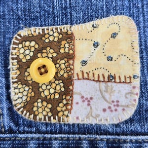May include: Close-up of a denim jacket with a decorative patch. The patch is rectangular with rounded corners and features a yellow button. The patch is made of four different patterned fabrics in shades of brown, yellow, and white, stitched together.