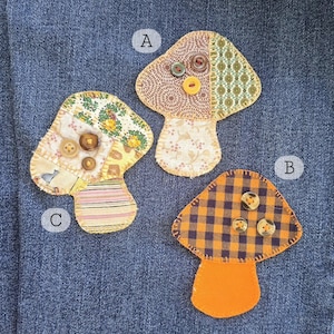 May include: Three handmade mushroom-shaped fabric patches. Each patch features a different patterned fabric in shades of brown, yellow, orange, and green. The patches are embellished with buttons and have a stem-like base.