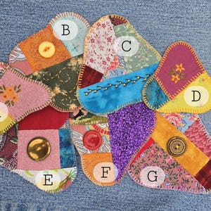 May include: A collection of colorful, handmade fabric coasters in various shapes and patterns. Each coaster features a unique patchwork design with different fabrics, colors, and textures, and is embellished with buttons. The coasters are arranged on a denim background.