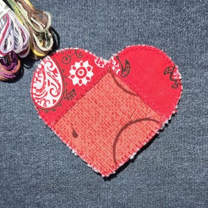 May include: A heart-shaped fabric patch in red, with paisley and floral patterns. The patch is made of several pieces of fabric stitched together. Embroidery floss in various colors is visible in the upper left corner. The patch is on a denim background.