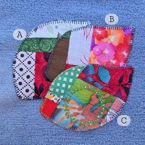 May include: Three colorful, leaf-shaped fabric patches with decorative stitching. Each patch is a patchwork of various patterned fabrics, including floral, polka dot, and geometric designs. The patches are arranged on a blue denim background.