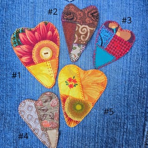 May include: Five heart-shaped fabric patches with unique designs and colors. Each patch is hand-stitched onto a denim background. The patches feature floral patterns, patchwork designs, and a button. The patches are numbered from #1 to #5.