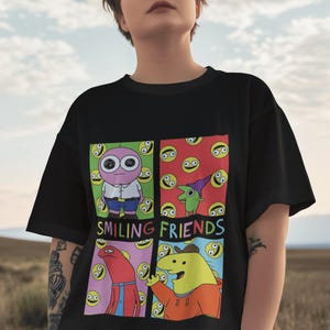 May include: Black t-shirt featuring a colorful graphic of cartoon characters in a grid pattern. The design includes the text "SMILING FRIENDS" in white letters. The characters are in various colors, including pink, green, red, and yellow.