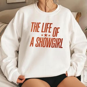 Könnte beinhalten: Weißes Sweatshirt mit dem Text "THE LIFE OF A SHOWGIRL" in glitzernden, orangefarbenen Buchstaben. Der Text ist zentriert und enthält drei kleine Sterne. Das Sweatshirt hat lange Ärmel und einen Rundhalsausschnitt.