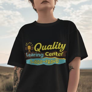 May include: Black t-shirt with the text "Quality Learing Center 1-800-FRAUD" in yellow, brown, and blue. The shirt has a graphic of a house with a tree on top. The shirt is a casual style.