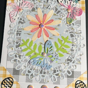 May include: Handmade Easter greeting card with a floral and butterfly theme. The card features a central flower, butterflies, and green leaves, with a decorative oval frame. The corners have Easter egg designs and a yellow and white checkered pattern.