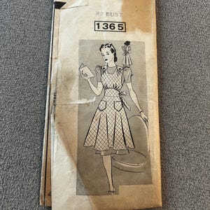 May include: A vintage paper sewing pattern envelope in tan with the text 42 BUST and 1365. The front features a black and white illustration of a person wearing a dress with a diamond-patterned apron, puff sleeves, and a tie waist, with a small inset showing the back view.
