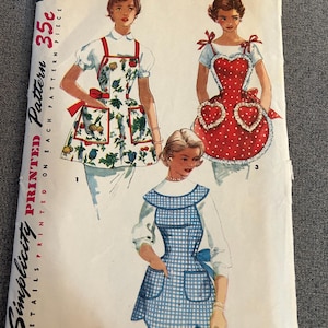 May include: Vintage Simplicity pattern envelope featuring illustrations of three aprons. The aprons include a floral print, a red polka dot heart design, and a blue gingham pattern. The envelope reads "ONE-YARD APRONS" and "Simplicity PRINTED Pattern 35c".