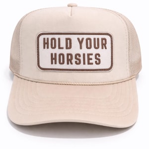 May include: A beige trucker cap with a mesh back and a curved peak. The front displays a rectangular patch with the words "HOLD YOUR HORSIES" in brown. A casual hat.